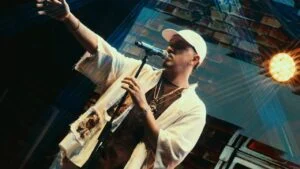 Delacruz canta no palco de camisa branca e boné, com o braço estendido