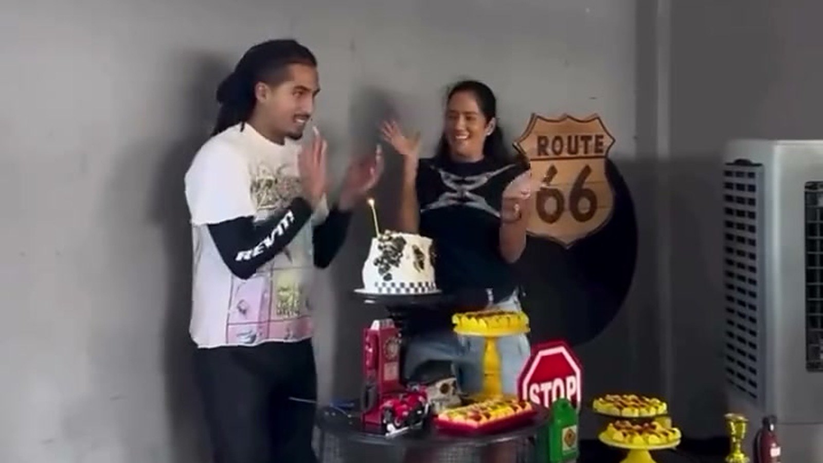 Matuê aplaude ao lado de uma mulher diante de um bolo com vela, em frente a uma placa Route 66